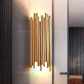 The Vigo Stainless Steel Wall Sconce by Morsale.com features vertical gold-toned rods with a radiant glow, mounted on a light wall in a modern, minimalist space.
