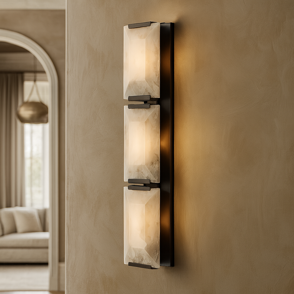 The Brookhill Calcite Crystal Sconce from Morsale.com, featuring three vertical frosted glass shades, emits a soft glow on a beige wall. An archway in the background leads to a bright living room.