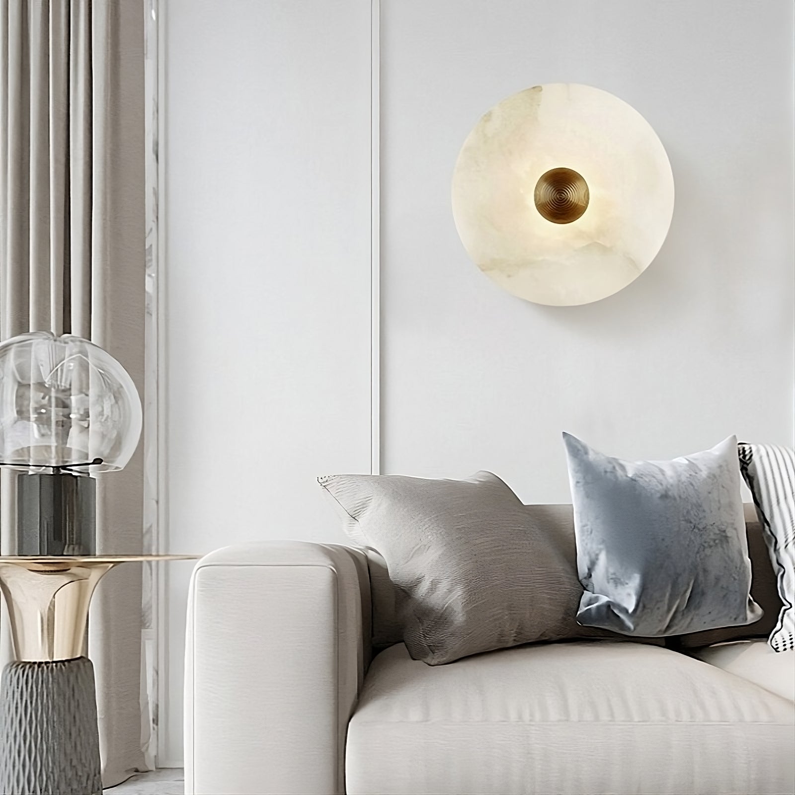 A minimalist living room with a modern design. A light gray sofa is adorned with gray and blue pillows. Mounted on the white wall, the Moonshade Natural Marble Wall Sconce from Morsale.com features a round, handmade design with a central gold accent, showcasing its unique texture. Beside the sofa, there is a small table with a glass lamp.