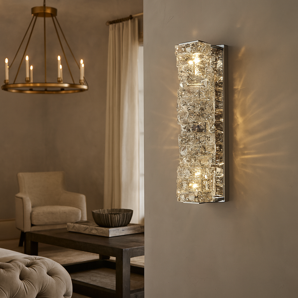 
                  
                    The Bacci Crystal Wall Sconce by Morsale.com features textured glass and chrome accents, casting intricate light patterns on a beige wall, while a brass chandelier glows above neutral furnishings in the cozy living area.
                  
                