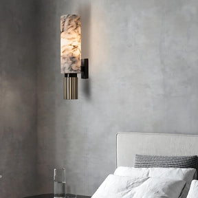 A Moonshade Natural Marble Sconce from Morsale.com, featuring a cylindrical marbled glass shade and a metallic base reminiscent of handmade marble sconces, is illuminated on a textured gray wall. Below it, a glass of water rests on a nightstand next to a gray upholstered bed adorned with white and black checkered pillows.