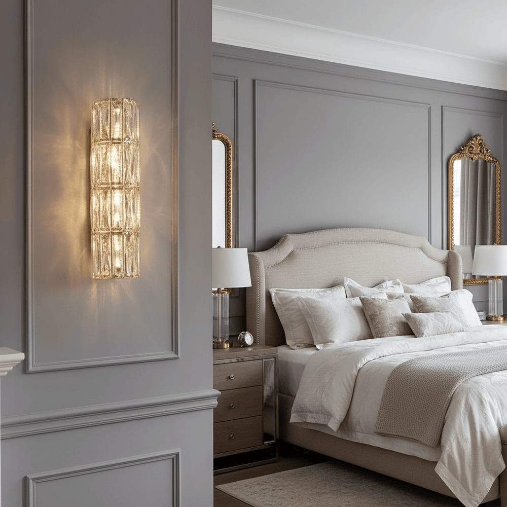 
                  
                    Elegant bedroom featuring a beige upholstered bed, white bedding, two bedside tables with lamps, ornate gold-framed mirrors, and the Bacci Crystal Sconce by Morsale.com casting a warm glow over gray paneled walls for modern flair.
                  
                