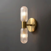 A Morsale Palermo 12" Brass Wall Light Sconce with two vertically aligned clear glass cylindrical shades, each patterned with a delicate diamond design. The base of the sconce is a circular brass mount attached to a dark grey wall, providing warm lighting that enhances its modern design.