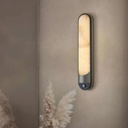 The Morsale Sandford Stainless Steel & Marble Wall Sconce, shaped like an elongated oval, offers a sophisticated wall accent with its warm glow. It stands against a neutral wall, while decorative pampas grass enhances its natural touch and complements the stainless steel elegance.