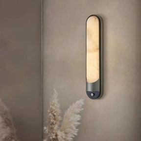 The Morsale Sandford Stainless Steel & Marble Wall Sconce, shaped like an elongated oval, offers a sophisticated wall accent with its warm glow. It stands against a neutral wall, while decorative pampas grass enhances its natural touch and complements the stainless steel elegance.