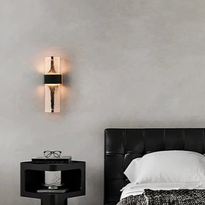 A modern bedroom with a black leather headboard bed, a white pillow, and a monochrome blanket features the sleek Water Ripple Crystal Wall Sconce by Morsale. The sconce casts a warm glow above a black bedside table holding glasses and a book against the gray wall, adding a touch of new classical style to the setting.