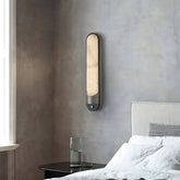 A modern bedroom features the Morsale Sandford Stainless Steel & Marble Wall Sconce, casting warm LED light on a gray textured wall near a white window. The serene scene is completed by a bed with white linens and a small glass of water on a dark nightstand.