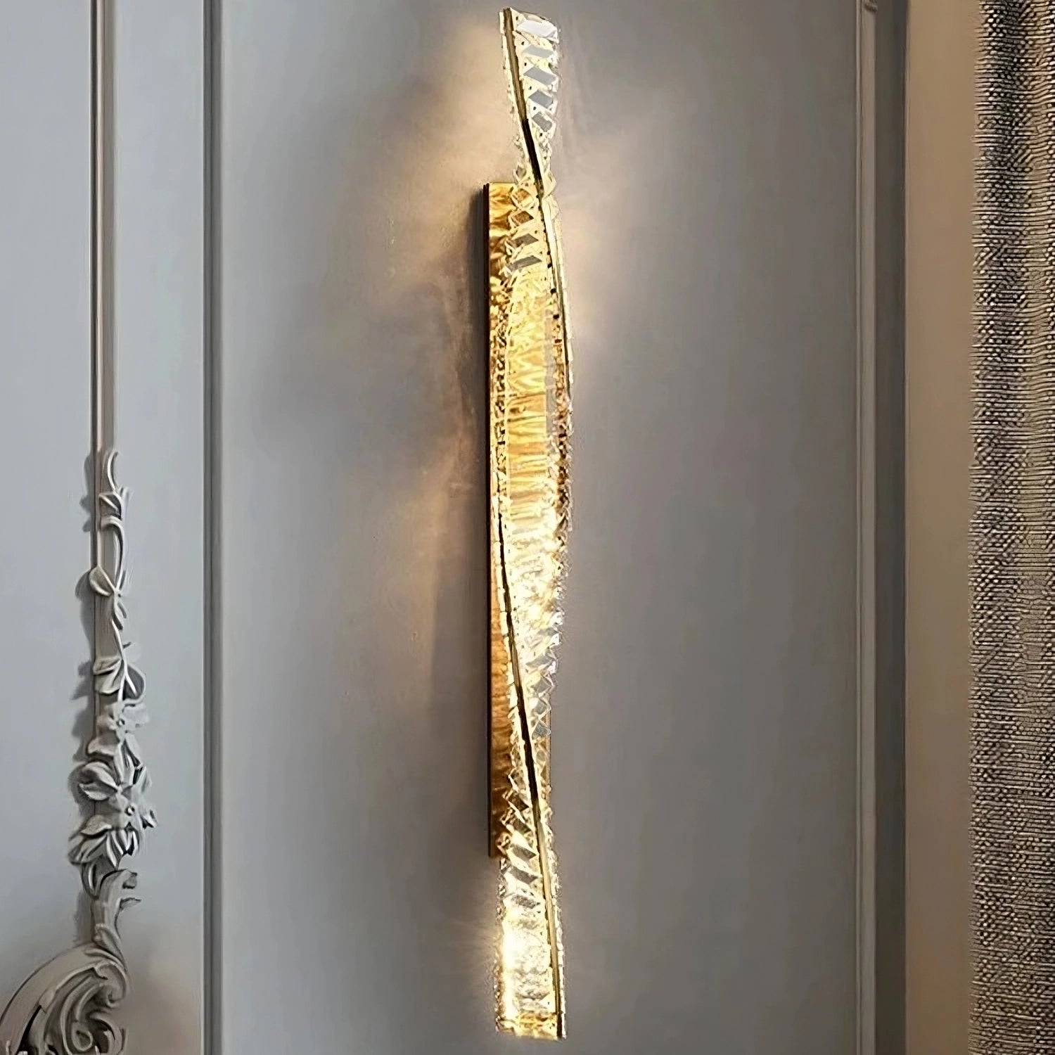 The Morsale Gio Crystal Sconce is a contemporary vertical wall lamp featuring a twisted metallic and glass design that casts a warm light. Its crystal accents add elegance, making it an ideal addition to sleek, minimalistic walls for enhancing the sophistication of interior decor.