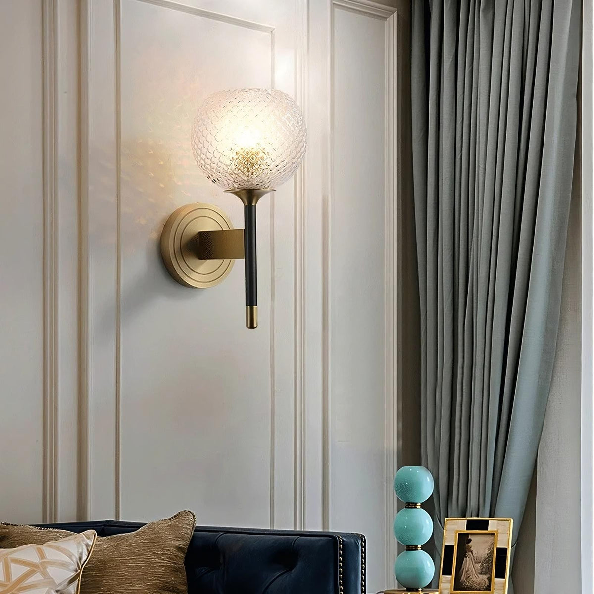 Modern living room featuring a sleek Morsale Palermo Wall Sconce Light with a textured glass globe and brass finish, mounted on a light gray paneled wall. Below, a navy tufted sofa with beige and patterned pillows sits beside a small table with blue stacked lamps and a framed photo.