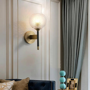 Modern living room featuring a sleek Morsale Palermo Wall Sconce Light with a textured glass globe and brass finish, mounted on a light gray paneled wall. Below, a navy tufted sofa with beige and patterned pillows sits beside a small table with blue stacked lamps and a framed photo.