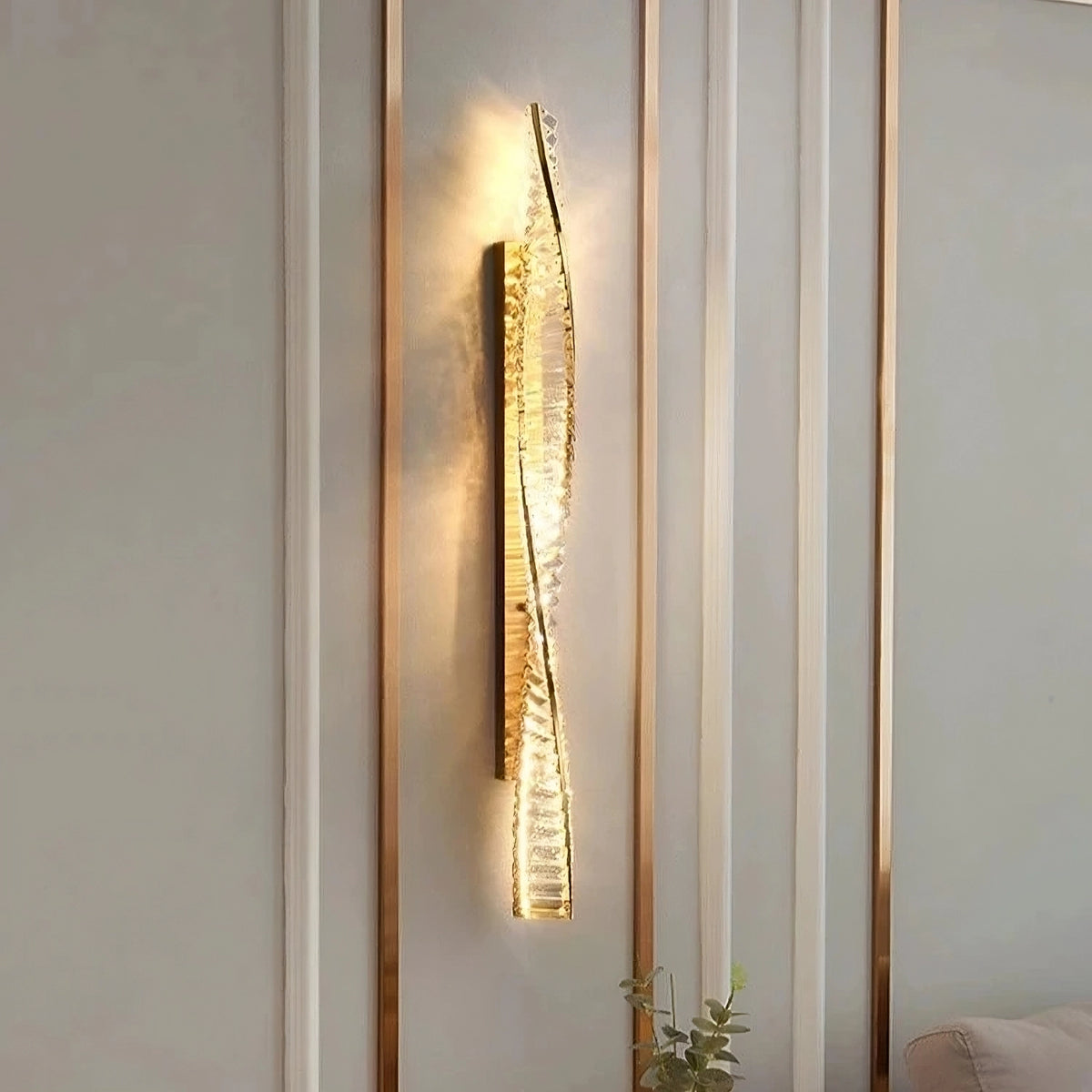 The Morsale Gio Crystal Sconce, featuring a modern twisted design and warm illumination, is mounted on a sleek wall adorned with crystal accents. This elegant fixture softly scatters light across the surface, enhancing the atmosphere.