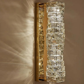 The Bacci Crystal Wall Sconce by Morsale.com, a gold wall-mounted light fixture, features a vertical arrangement of handmade crystals, creating a visually striking pattern. Equipped with E14 Base LED Bulbs, it emits a warm, ambient glow reflected on the adjacent wall.