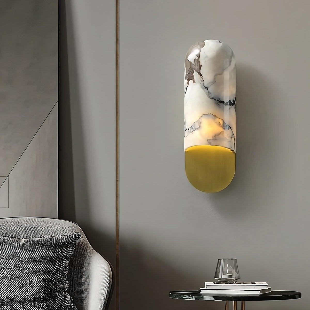 A Natural Marble Wall Sconce Light Fixture by Morsale.com hangs on a sleek gray wall above a small round side table with a glass of water. Part of a gray armchair with textured fabric is visible in the foreground. The room features minimalist and contemporary decor.
