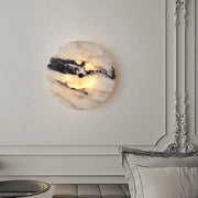 A modern interior showcases a Morsale Moonshade Natural Marble Sconce Light, wall-mounted and energy-efficient, with distinct black and white patterns emitting a soft glow. It's positioned on an intricately paneled wall above a gray cushioned bench beside a partially visible gray pillow.
