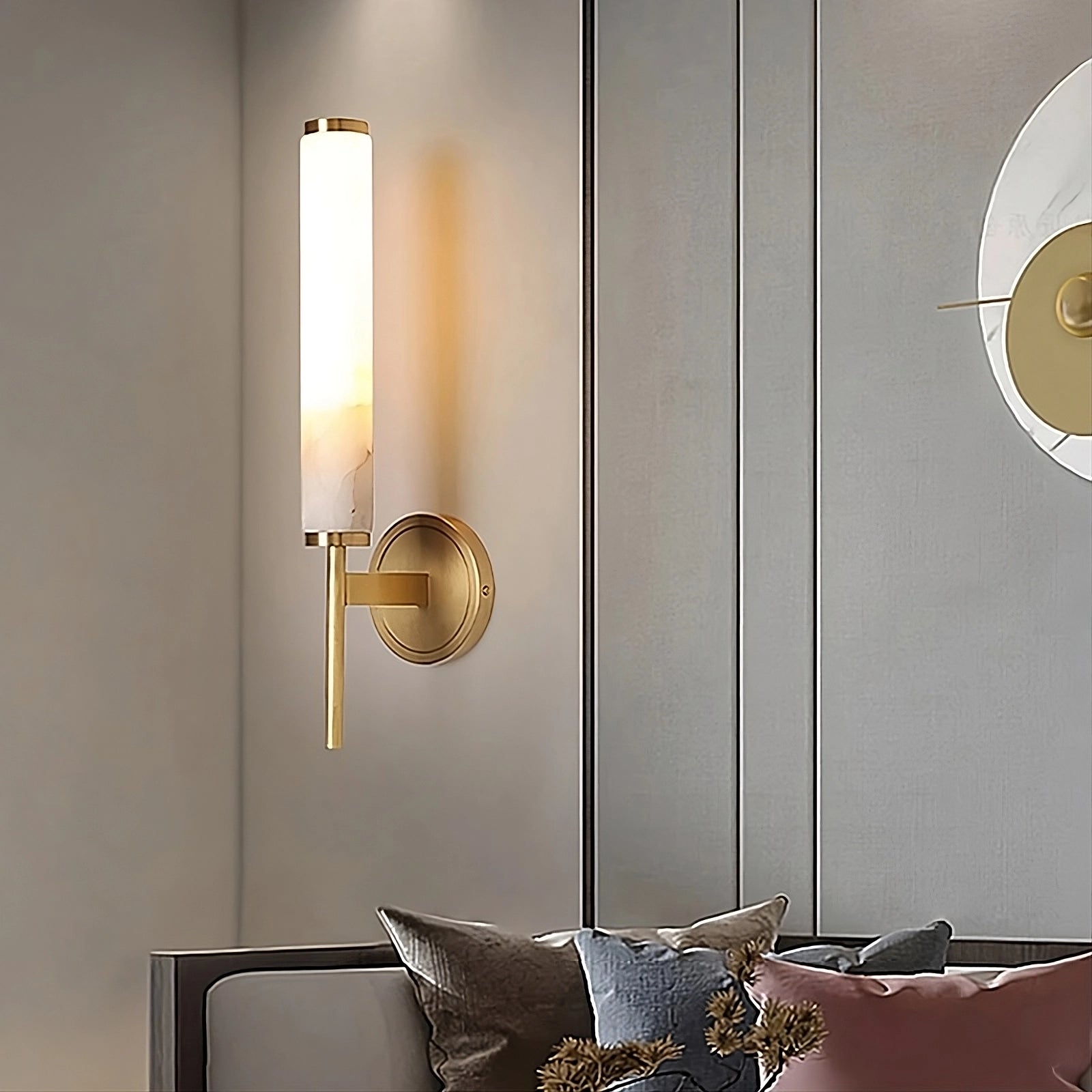 Mounted on a gray paneled wall is Morsale.com's Natural Marble & Copper Candle Style Wall Sconce, featuring a sleek gold base and a cylindrical white shade. This modern, minimalistic luxury home decor piece illuminates the sofa section below, where cushions in various colors like gray and pink are adorned with gold embellishments.