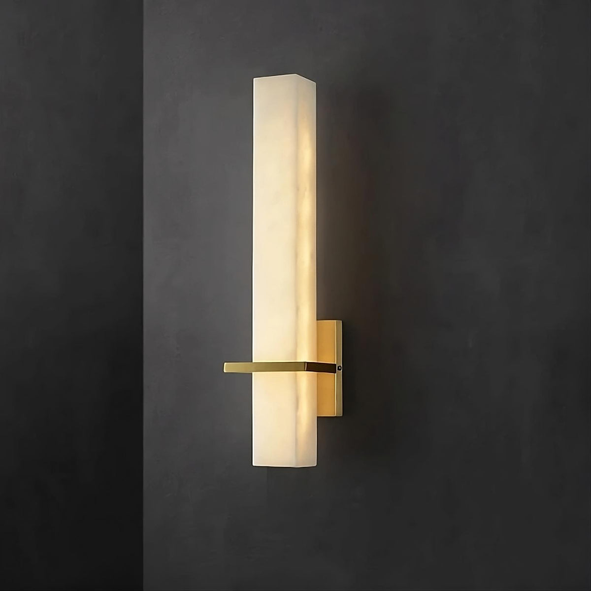 A modern Natural Marble Indoor Sconce with a sleek, vertical rectangular design emits a soft, ambient light. Featuring a frosted glass shade and matte gold metal base, this Morsale.com LED light fixture is mounted on a dark charcoal gray wall. The minimalist design adds an elegant touch to the setting.