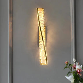 The Morsale Bacci Crystal Wall Sconce, featuring a twisted crystal-like design, casts warm ambient light on a gray wall. A vase with white and pink flowers is partially visible in the lower right corner.
