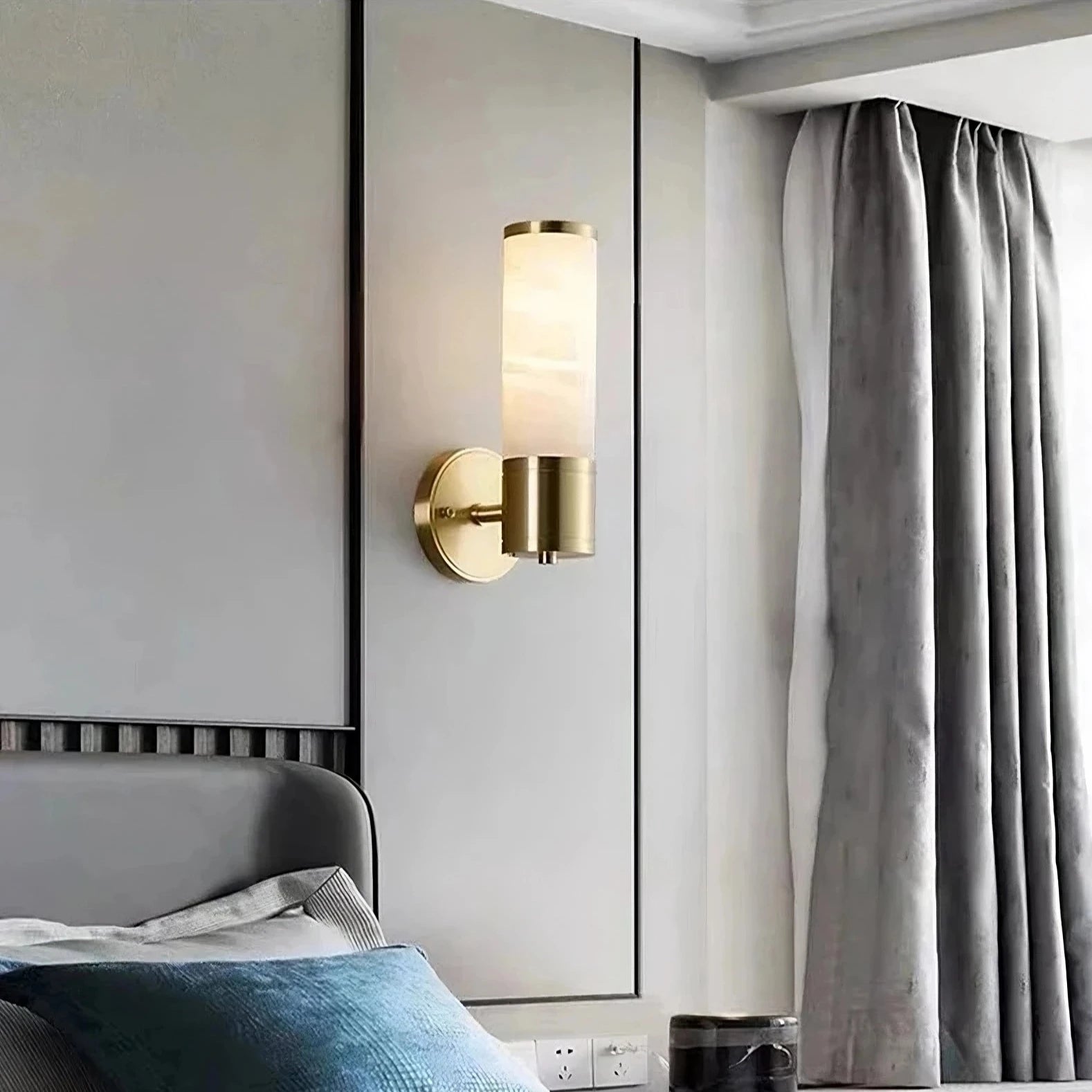 A modern bedroom features Moonshade Natural Marble Sconce Lights from Morsale.com, with genuine marble and frosted glass, emitting a warm light above a bed with a gray headboard. The room has light gray paneled walls and large floor-to-ceiling curtains in gray framing a window. The bed is adorned with blue and gray pillows.