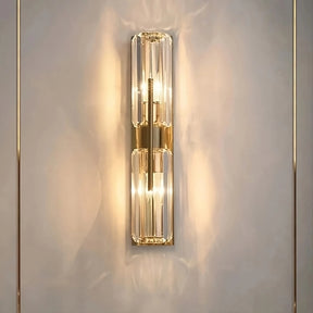 The Gio Double Head Crystal Wall Sconce by Morsale features two elongated, clear glass light fixtures on a metallic base that emit a warm and soft yellow glow against a light-colored wall. This modern LED wall lamp enhances elegance and creates a cozy ambiance.