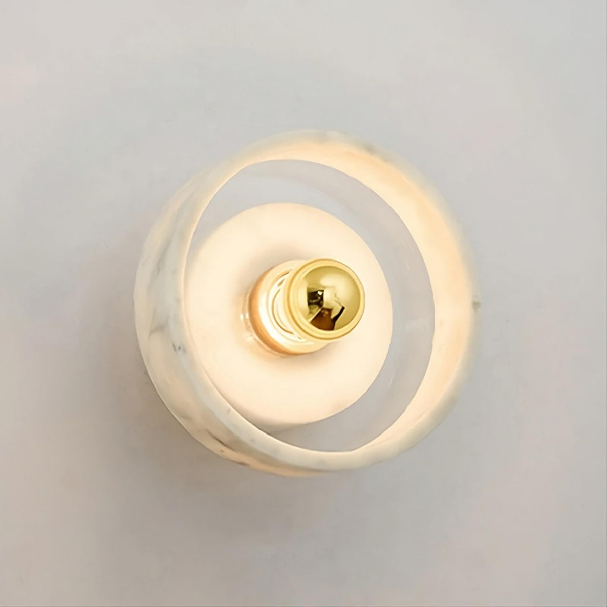 A modern wall light fixture with a minimalist design features a central light bulb surrounded by a circular, translucent white ring. The soft light emitted creates a warm ambiance on a plain, light-colored background, enhanced by the unique texture of the Moonshade Natural Marble Wall Light Fixture from Morsale.com.