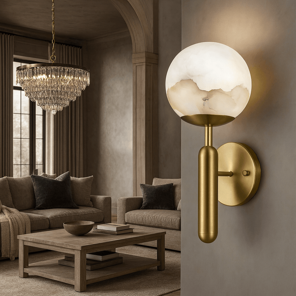 A modern living room features the Orla Marble Sphere Wall Sconce by Morsale.com in the foreground, with a crystal chandelier, neutral sofa, and wooden coffee table with a bowl completing the elegant scene.