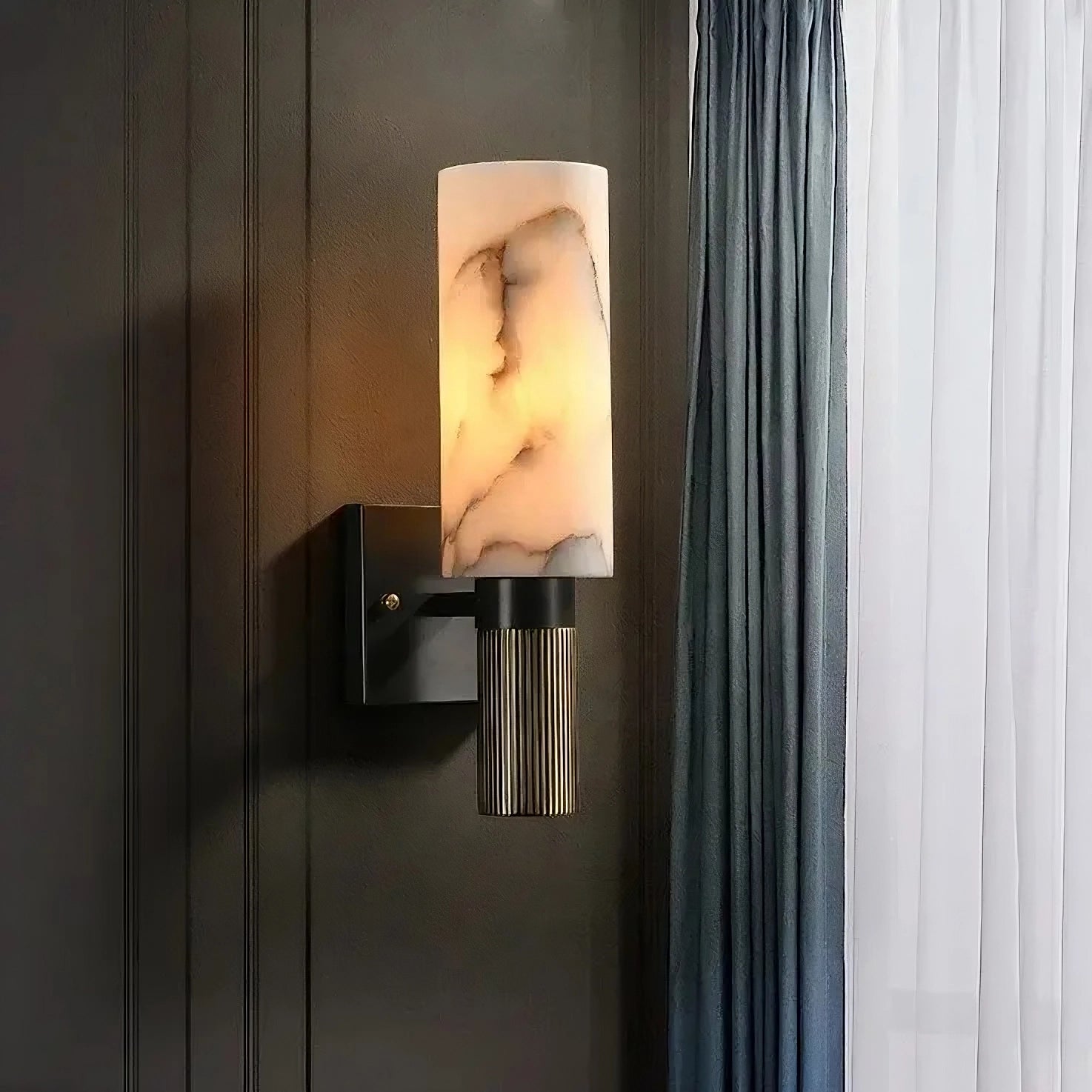 The Moonshade Natural Marble Sconce by Morsale.com is a modern wall fixture featuring a cylindrical, premium natural marble lampshade and a ribbed metallic base mounted on a dark rectangular bracket. It emits a warm glow, lighting up part of the textured wall, with blue and white curtains partially visible to the right.