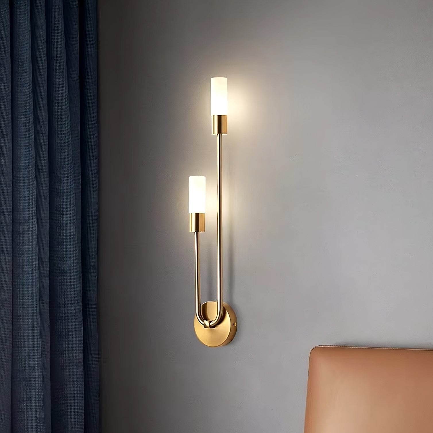 The Morsale Palermo 2-Light LED Wall Sconces, featuring two vertical cylindrical LED lights on a gold-colored base, are installed on a neutral wall. This energy-efficient fixture is enhanced by the presence of dark curtains and a glimpse of a beige sofa in the background, establishing an elegant ambiance.