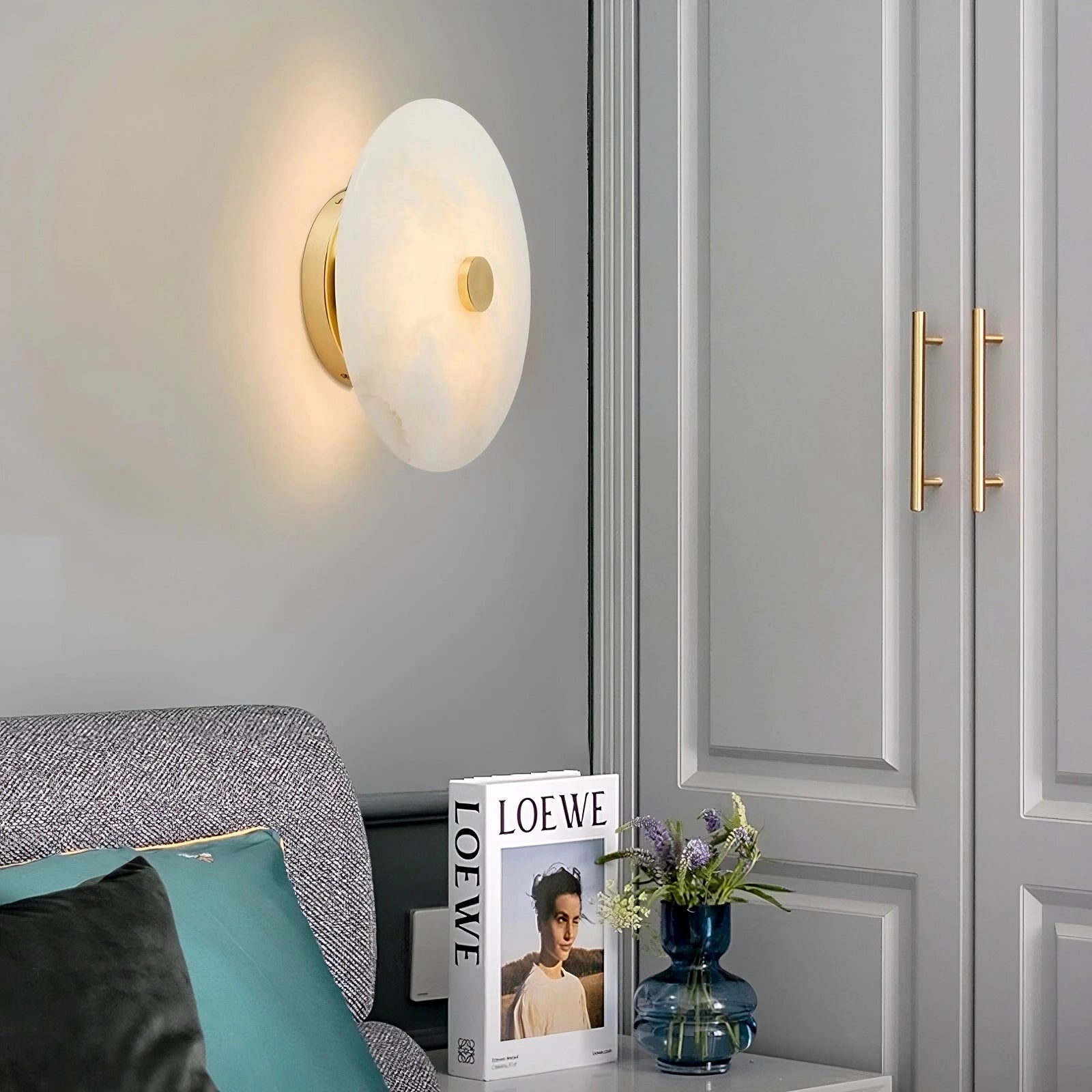 A modern living room corner features the Morsale Witney Copper & Marble Wall Sconce, a gray couch with teal and gray pillows, a side table displaying "LOEWE" and a bouquet in a blue vase, against refined gray paneled cabinets.