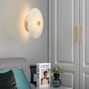 A modern living room corner features the Morsale Witney Copper & Marble Wall Sconce, a gray couch with teal and gray pillows, a side table displaying "LOEWE" and a bouquet in a blue vase, against refined gray paneled cabinets.