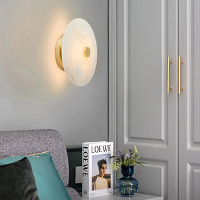 A modern living room corner features the Morsale Witney Copper & Marble Wall Sconce, a gray couch with teal and gray pillows, a side table displaying "LOEWE" and a bouquet in a blue vase, against refined gray paneled cabinets.