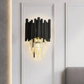 The Vigo Crystal Wall Sconce by Morsale.com features black and gold cylindrical tubes, E14 LED bulbs, and warm light. Shown mounted on a white paneled wall above a gray upholstered surface.