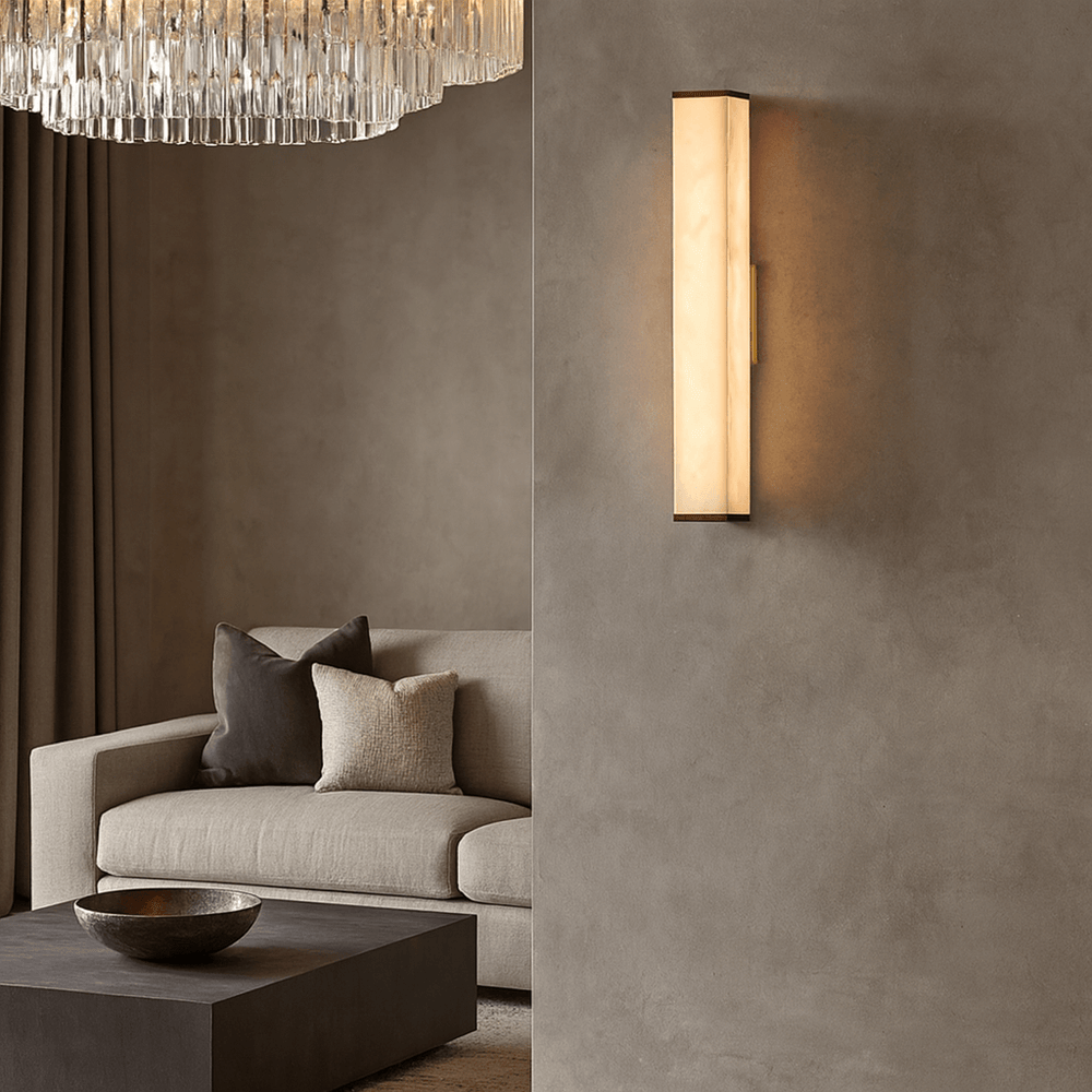 A modern living room features a beige sofa, neutral pillows, a dark coffee table with a decorative bowl, and the Avana Marble Wall Sconce by Morsale.com casting warm light on a textured wall beneath a crystal chandelier.