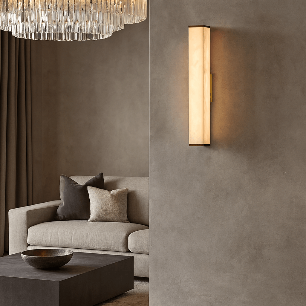 A modern living room features a beige sofa, neutral pillows, a dark coffee table with a decorative bowl, and the Avana Marble Wall Sconce by Morsale.com casting warm light on a textured wall beneath a crystal chandelier.