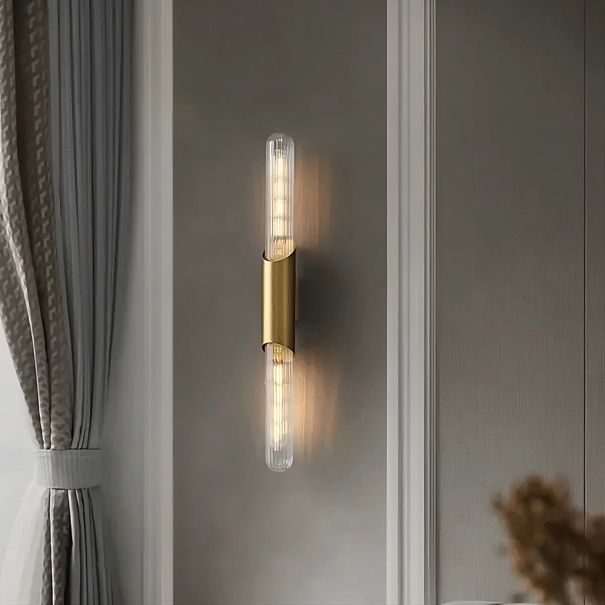 A Morsale Palermo 23" Brass Wall Sconce Light with two tubular bulbs is mounted on a light gray wall, exuding elegant lighting. To the left, a window with gray curtains partially draped by white sheers adds to the sophisticated ambiance of the space.