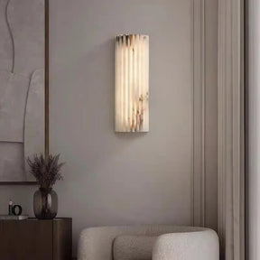 A cozy living room features a textured white armchair with Morsale's Natural Marble LED Wall Sconce, casting soft light on framed abstract art. A small wooden table with a vase of dried flowers completes the inviting atmosphere.