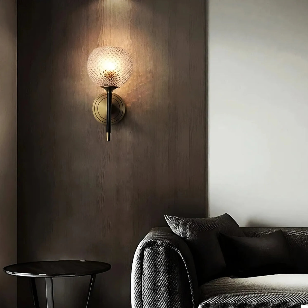 A modern living room with a black sectional couch and dark cushions. A round black side table is next to the couch. The Morsale Palermo Wall Sconce Light, featuring a textured glass globe and brass finish, emits a warm light on the dark paneled wall, creating a cozy ambiance.