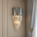 The Gio Crystal & Steel Sconce by Morsale.com features a geometric metal frame and hanging crystal prisms, casting warm light on a beige paneled wall beside gray curtains with a gold tieback.