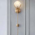 A minimalist, gold Morsale.com Natural Marble Wall Light Sconce with a round marble shade featuring unique marble patterns is mounted on a light gray, paneled wall. The sconce has a slender, vertical rod that extends downward from the circular base to a small, tapered end. Soft, warm light emits from the genuine marble lighting shade.