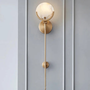 A minimalist, gold Morsale.com Natural Marble Wall Light Sconce with a round marble shade featuring unique marble patterns is mounted on a light gray, paneled wall. The sconce has a slender, vertical rod that extends downward from the circular base to a small, tapered end. Soft, warm light emits from the genuine marble lighting shade.