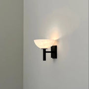 The Morsale Spanish Marble & Copper Antique Sconce, featuring a black mounting fixture and a white, semi-circular shade, casts a soft, warm glow that gently highlights a minimalist white wall while incorporating an elegant hint of copper antique sophistication.