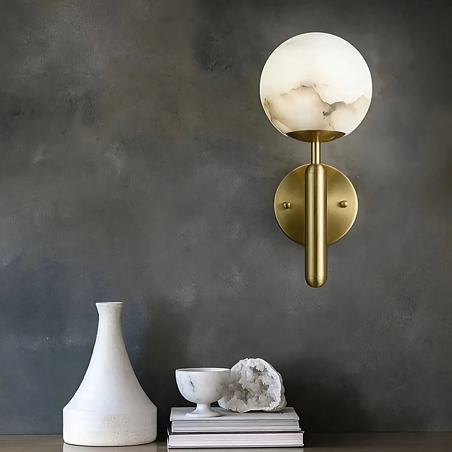 A modern wall sconce with a brass base and frosted glass globe is mounted on a dark, textured wall. Below it, a minimalist white vase, a white bowl, a crystal, and a small stack of books are neatly arranged on a table or shelf. The Natural Marble Sphere Wall Sconce by Morsale.com adds an elegant touch to the setup.
