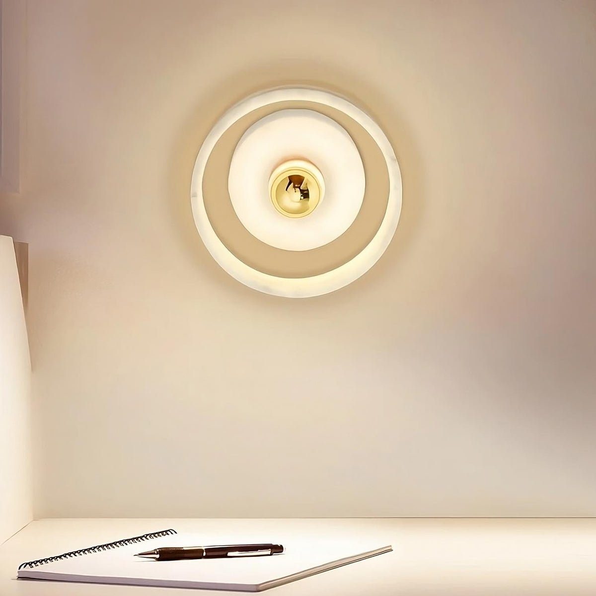 A modern Morsale.com Moonshade Natural Marble Wall Light Fixture with concentric circular designs illuminates a minimalistic workspace. Below, a white desk holds a black pen and an open spiral notebook, creating a clean and focused environment.