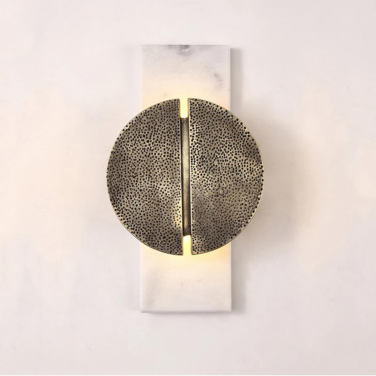 A modern wall sconce with a circular, textured metal design, split symmetrically down the middle. The light emits softly from behind, mounted on a rectangular base against a plain, light-colored wall. The Medieval Marble Wall Light Sconce by Bigman adds an elegant touch to any space.