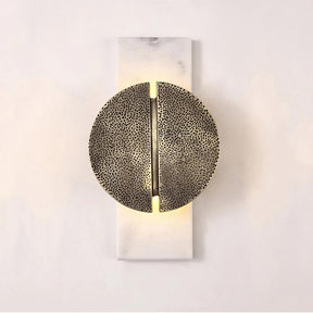 A modern wall sconce with a circular, textured metal design, split symmetrically down the middle. The light emits softly from behind, mounted on a rectangular base against a plain, light-colored wall. The Medieval Marble Wall Light Sconce by Bigman adds an elegant touch to any space.