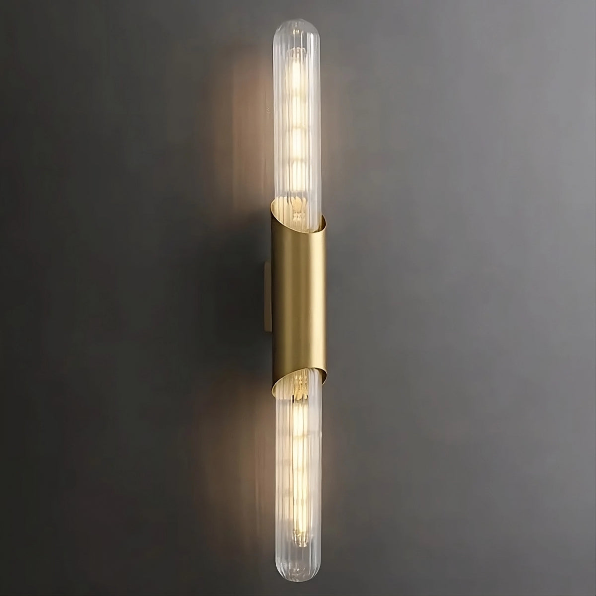 A modern wall sconce with a vertical, cylindrical design features two elongated, clear glass bulbs on either end. The bulbs are encased in a sleek brass holder, emitting warm light against a dark gray background, exemplifying elegant lighting with the Palermo 23" Brass Wall Sconce Light by Morsale.