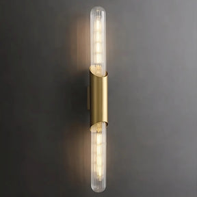 A modern wall sconce with a vertical, cylindrical design features two elongated, clear glass bulbs on either end. The bulbs are encased in a sleek brass holder, emitting warm light against a dark gray background, exemplifying elegant lighting with the Palermo 23" Brass Wall Sconce Light by Morsale.