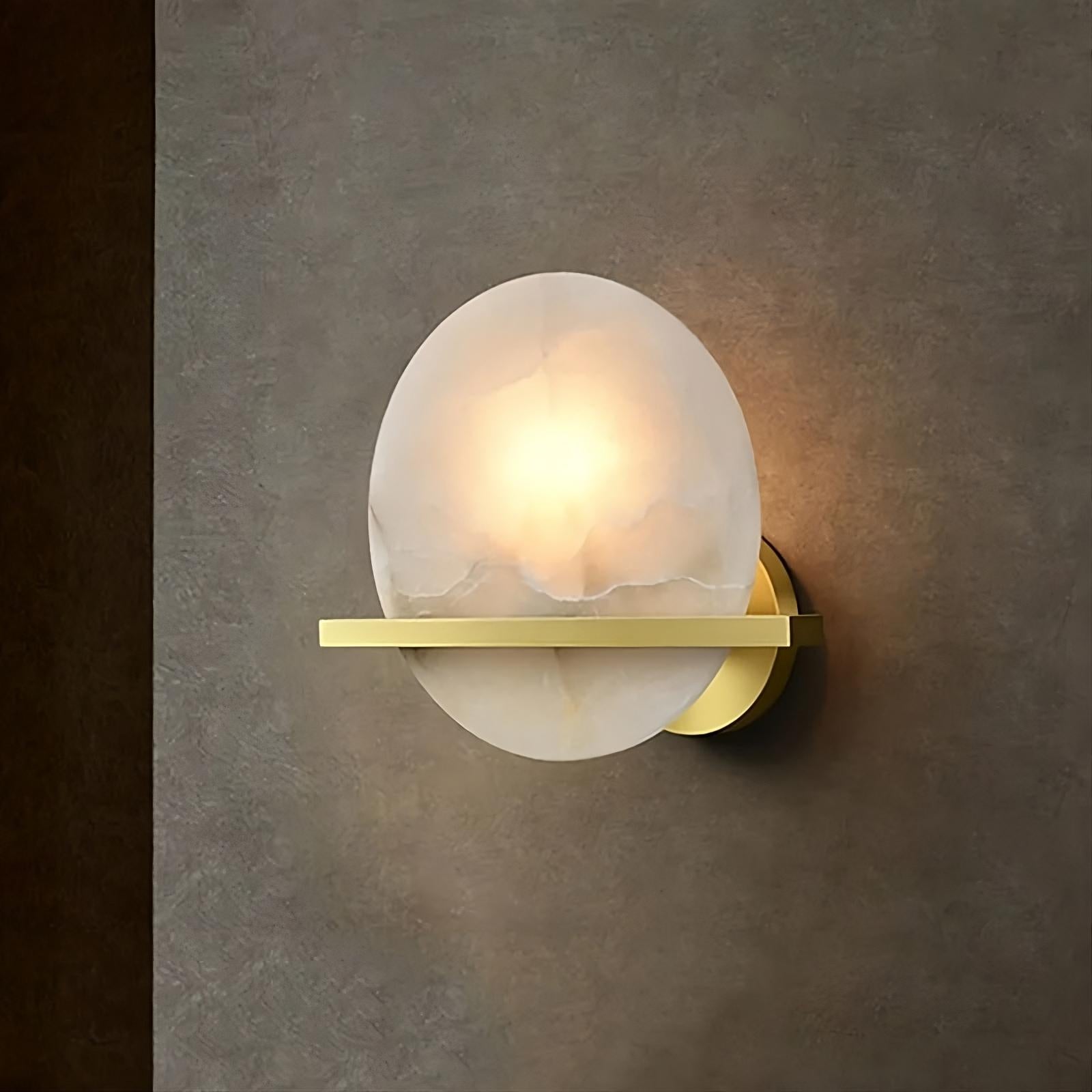 A modern wall sconce with a circular, frosted white glass shade emits a warm LED light source. The fixture is mounted on a smooth, dark wall and features a sleek, horizontal gold accent bar that appears to divide the sconce, embodying contemporary design. This exquisite piece is the Natural Marble Wall Light Sconce by Morsale.com.