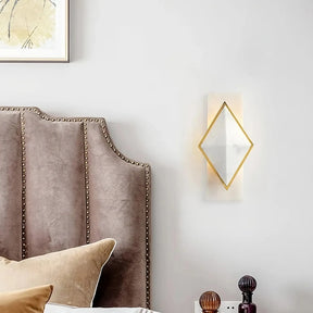 A cozy bedroom features a plush bed with a tufted headboard, neutral-toned pillows, and the Natural Marble Contemporary Wall Sconce Light by Bigman. The light's art deco charm and gold frame cast a warm glow. A framed abstract artwork hangs above the bed, complemented by a small bedside table adorned with decorative items.