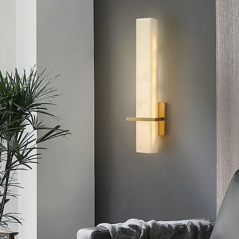 A modern Natural Marble & Brass Wall Sconce from Morsale.com with a rectangular, frosted glass shade and a gold metal mount is illuminated, casting a soft glow. It is mounted on a dark-colored wall adjacent to a white wall. A plant and part of an elegant sofa with a blanket are visible in the room.