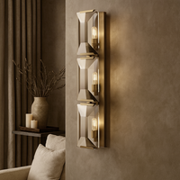 The Brookhill Crystal Wall Sconce by Morsale.com features three geometric glass panels and warm bulbs on a taupe wall, accented by beige curtains, a branch vase, candles, and a cream sofa.
